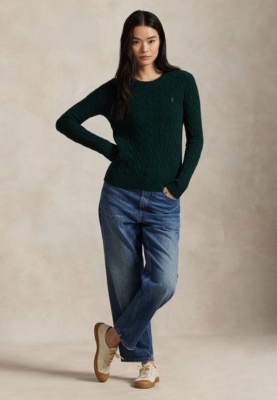 Donna con capelli lunghi e scuri indossa un maglione a trecce verde scuro, jeans blu e sneakers beige, in piedi con una mano sul fianco e l'altra in tasca.