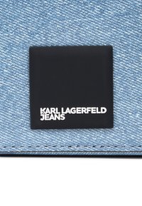 Denimový materiál s modrým tkaním a čiernou obdĺžnikovou etiketou. Na etike je napísané "KARL LAGERFELD" a "JEANS" bielym písmom.