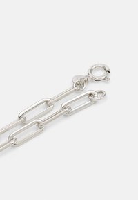 Silver kedja armband med förlängda länkar, en krabba lås och en liten charm. Ytan är slät och glänsande.