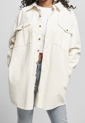 Personne portant une chemise boutonnée oversize écrue sur un crop top blanc et un jean bleu clair sur un fond uni.