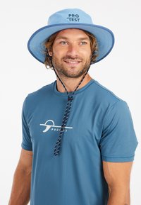 Lichtblauwe brede randemmer met marineblauwe afwerking en verstelbare kinriem. Draagt een blauwe gemêleerde t-shirt met een wit logo.