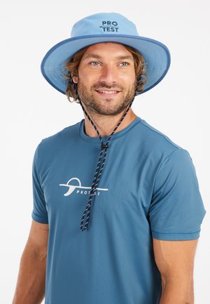 Light blue wide-brimmed bucket hat with navy trim and adjustable chin strap. Worn with a blue textured t-shirt featuring a white logo.