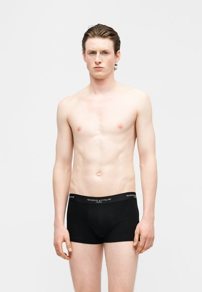 Νέος άνδρας με ανοιχτόχρωμο δέρμα και κοντά καστανά μαλλιά, φοράει μαύρα μποξεράκια Maison Kitsuné, στέκεται μπροστά σε λευκό φόντο.