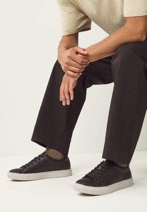 Homme assis, mains jointes, portant une chemise beige, un pantalon noir, des baskets marron foncé et des chaussettes marron sur un fond uni.