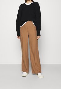 Donna che indossa un maglione nero sopra una camicia bianca, pantaloni larghi marroni e sneakers bianche, in piedi contro uno sfondo chiaro e uniforme.