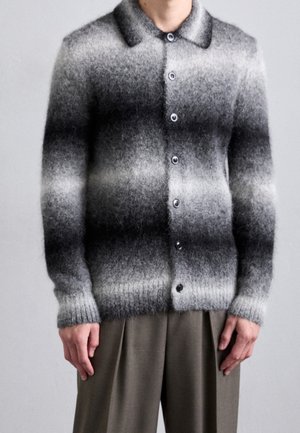 Mężczyzna ubrany w zapinany na guziki, miękki sweter rozpinany z poziomymi, szaro-czarnymi paskami gradientowymi, w połączeniu z luźnymi oliwkowozielonymi spodniami.