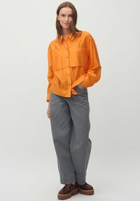 Camicetta arancione con bottoni, dal taglio rilassato e due tasche sul petto, abbinata a pantaloni grigi a gamba larga e scarpe platform marroni scure.