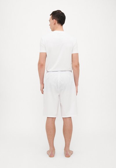 Weißes T-Shirt mit kurzen Ärmeln und passende Shorts, beide aus glattem Stoff, von hinten auf einem barfüßigen männlichen Modell vor einem schlichten Hintergrund präsentiert.