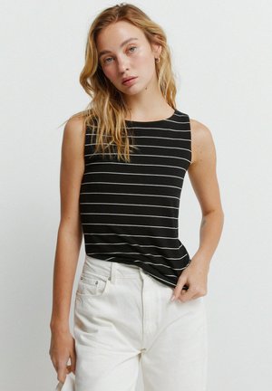 Jonge vrouw met blond haar, gekleed in een zwart mouwloos shirt met witte horizontale strepen en witte broek met hoge taille, voor een effen achtergrond.