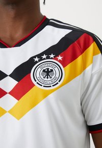 Gros plan sur un maillot de football allemand blanc avec des rayures diagonales noires, rouges et jaunes, et le logo Deutscher Fussball-Bund orné de quatre étoiles.