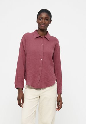 Kvinne iført en langermet, texturert mauve skjorte med knapper og lysebeige bukser, står mot en enkel hvit bakgrunn.