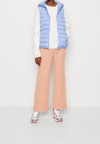 Jasnoniebieska kamizelka puchowa z kapturem na białym swetrze, zestawiona z brzoskwiniowymi szerokimi spodniami w prążki i sneakersami z srebrnymi detalami.