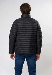 Schwarze Puffjacke mit horizontalen Nähten, hohem Kragen und glatter Textur. Getragen mit blauen Jeans, die die Rückansicht zeigt.