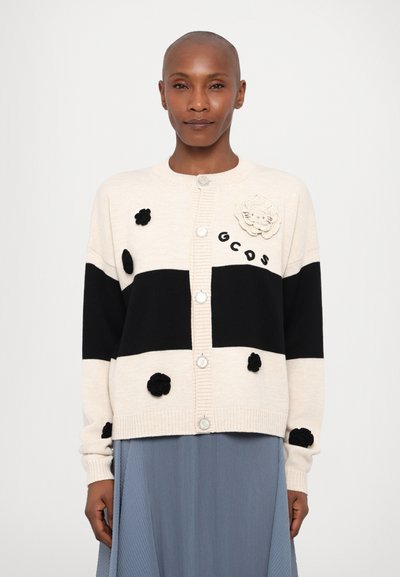 Strikket cardigan i beige og sort med runde sølvknapper, dekorativ blomsterapplikation og teksturerede sorte pom-poms på fronten.