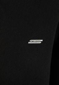 T-shirt noir ajusté avec une texture lisse, présentant une petite étiquette métallique en argent portant la mention "SMILODOX" sur le devant.