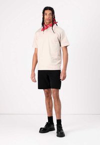 Lys beige bomull t-skjorte, korte ermer, kombinert med svarte denimshorts. Modellen har på seg et rødt bandana og svarte platå sko.