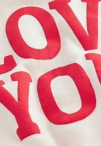 Tela blanca con letras rojas en negrita que dicen "TE AMO." La textura es suave con puntos de tejido visibles de la prenda.