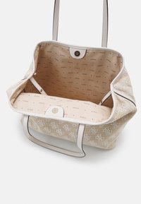 Sac cabas tissé beige avec poignées en cuir blanc et doublure intérieure en tissu imprimée du motif du logo "GUESS". Fermeture pression magnétique visible.