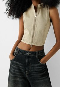 Beige cropped denimvest med høj krave og lynlås foran, kombineret med mørkeblå viddeben-jeans med kontrastsyning og lommer.