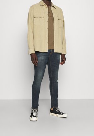 Camisa de pana beige con dos bolsillos en el pecho, polo marrón debajo, pantalones ajustados azul oscuro y zapatillas altas negras con suelas blancas.