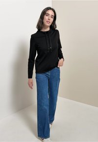 Maglietta nera a maniche lunghe con cappuccio e cordini, abbinata a jeans denim blu larghi. Il modello si trova di fronte a uno sfondo neutro.