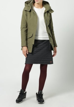 Wodoodporna kurtka w oliwkowej zieleni z kapturem, zestawiona z czarną pikowaną spódnicą i bordowymi rajstopami; czarne buty sportowe dopełniają strój.