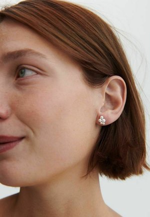 Noorpruunide lühikeste juustega noor naine kannab vasakul kõrvasiblal viiest väikesest valgest pärlikõrvarõngast koosnevat komplekti, vaadates vasakule ja kerge naeratusega.