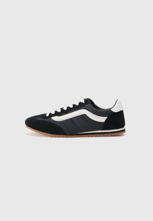 Sort-hvid lavtop sneakers med hvide snørebånd, en hvid sidelinje, brun gummisål og hvid hælkappe på en ensfarvet lys baggrund.
