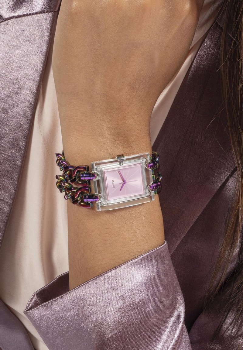 Montre carrée avec un boîtier transparent, un cadran rose, un bracelet en maillons de chaîne violet et des éléments en argent. Portée sur un poignet contre un fond satiné.