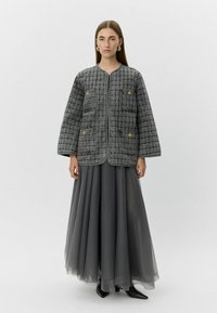 Chaqueta gris acolchada con cuatro bolsillos frontales, escote redondo y detalles de botones dorados, combinada con una falda larga y fluida gris.