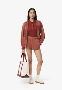 Burgundy huppari, vastaava polo ja pehmeästä kankaasta valmistetut shortsit; vaaleanruskea kassi burgundy-yksityiskohdilla; valkoiset lenkkarit beige-pohjalla.