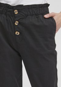 Pantalons noirs avec une taille élastique, comportant trois boutons beiges sur le devant et des poches latérales. Tissu lisse avec une finition mate.