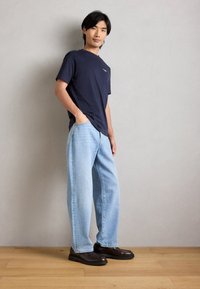 Μαύρη μπλούζα crew-neck, ανοιχτό μπλε παντελόνι με φαρδιά ποδιά, και καφέ δερμάτινα παπούτσια. Το μοντέλο στέκεται με τα χέρια στις τσέπες, μπροστά από ουδέτερο φόντο.