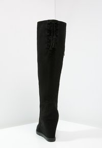 Bota negra por encima de la rodilla hecha de ante, con un tacón en cuña, detalles de encaje en la parte trasera y una textura suave.