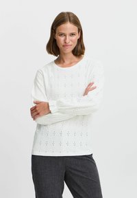 Maglietta bianca a maniche lunghe con design a occhio, scollatura rotonda e polsini elasticizzati, abbinata a pantaloni grigio scuro. Materiale testurizzato e leggero.