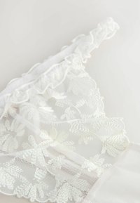 White lace bralette featuring floral embroidery, ruffled trim, and sheer mesh panels, with a smooth back layer and soft texture.