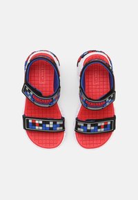 Punaiset sandaalit, joissa on mustat ja siniset säädettävät hihnat, joissa on teksturoitu pohja ja ruutukuvioiset yksityiskohdat hihnoissa. Kevyt muotoilu.