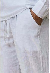 Chemise en lin blanc à coupe ample, avec des manches longues et un cordon de serrage à la taille, présentant un tissu légèrement texturé.