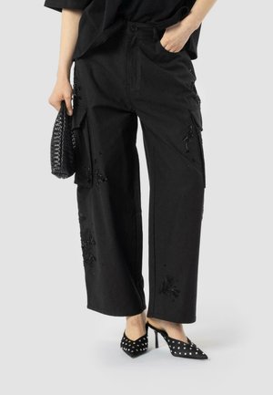Black cargo trousers with decorative embroidery, wide leg design, paired with black studded heels and a textured clutch.