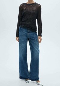 Zwarte gebreide langslijf top met een losse weving, gecombineerd met wijde blauwe jeans. Zilveren puntige hakken zorgen voor een stijlvolle touch.