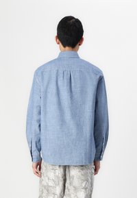 Knapp-up skjorte i lyseblå chambray stof, lange ærmer, enkelt plissé detalje bagpå, parret med mønstrede, lyse bukser.