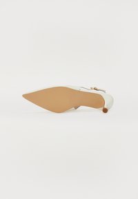 Chaussure à talon haut blanche avec un bout pointu et une semelle beige. Présente un petit nœud décoratif et un accent près du talon. Texture lisse.