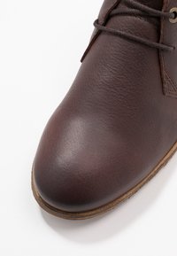 Stivaletto in pelle marrone con una texture liscia, punta tonda e suola in gomma chiara. Dettagli di cucitura sottili visibili.