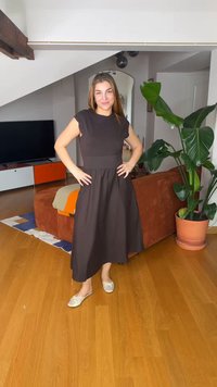 Γυναίκα που φοράει μαύρο μεσαίο φόρεμα και κρεμ φλατ παπούτσια, στέκεται με τα χέρια στη μέση σε ένα μοντέρνο σαλόνι με καφέ καναπέ και μεγάλο πράσινο φυτό.