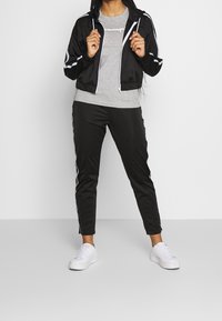 Svart zip-up jacka med vita ränder, grå t-shirt med logotyp och svarta smalrandiga byxor, kombinerat med vita sneakers.