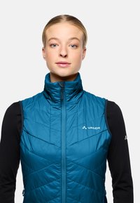 Türkise, isolierte Weste mit gestepptem Design, hohem Kragen und Vorderreißverschluss. Mit Logo auf der linken Brust und strapazierfähigem Material.