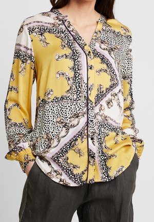Kvinde iført en langærmet knap-op bluse med et gult, sort, hvidt og lilla barok- og leopardprint-mønster, med hænderne i lommerne.