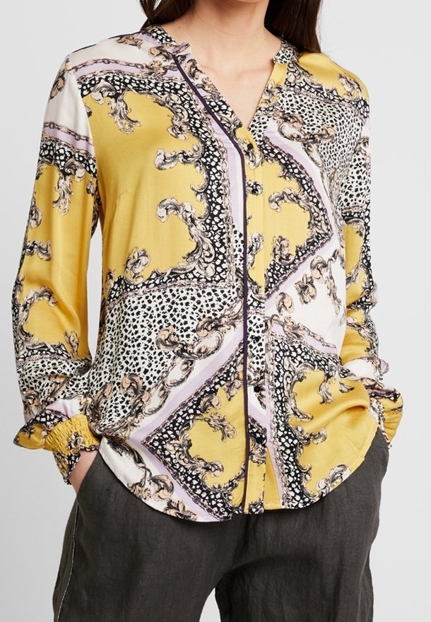 Femme portant un chemisier à manches longues boutonné avec un motif baroque et léopard jaune, noir, blanc et violet, les mains dans les poches.