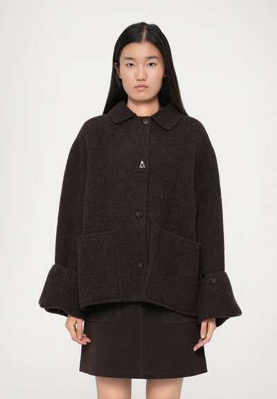 Young woman with long straight hair wearing a dark brown wool coat with wide cuffs and large front pockets over a matching skirt.