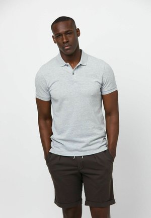 Camiseta polo gris de manga corta con cuello y tres botones, combinada con pantalones cortos oscuros enrollados, posando contra un fondo blanco.